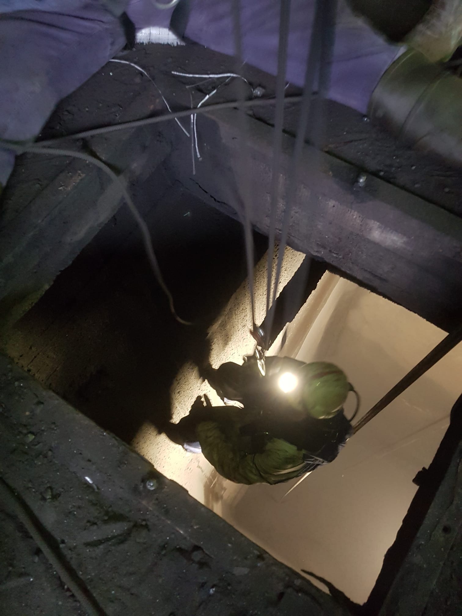 Pracownik na linie, oświetlony reflektorem, opuszcza się do ciemnego otworu w dachu. Widoczne liny asekuracyjne i fragment konstrukcji dachu.