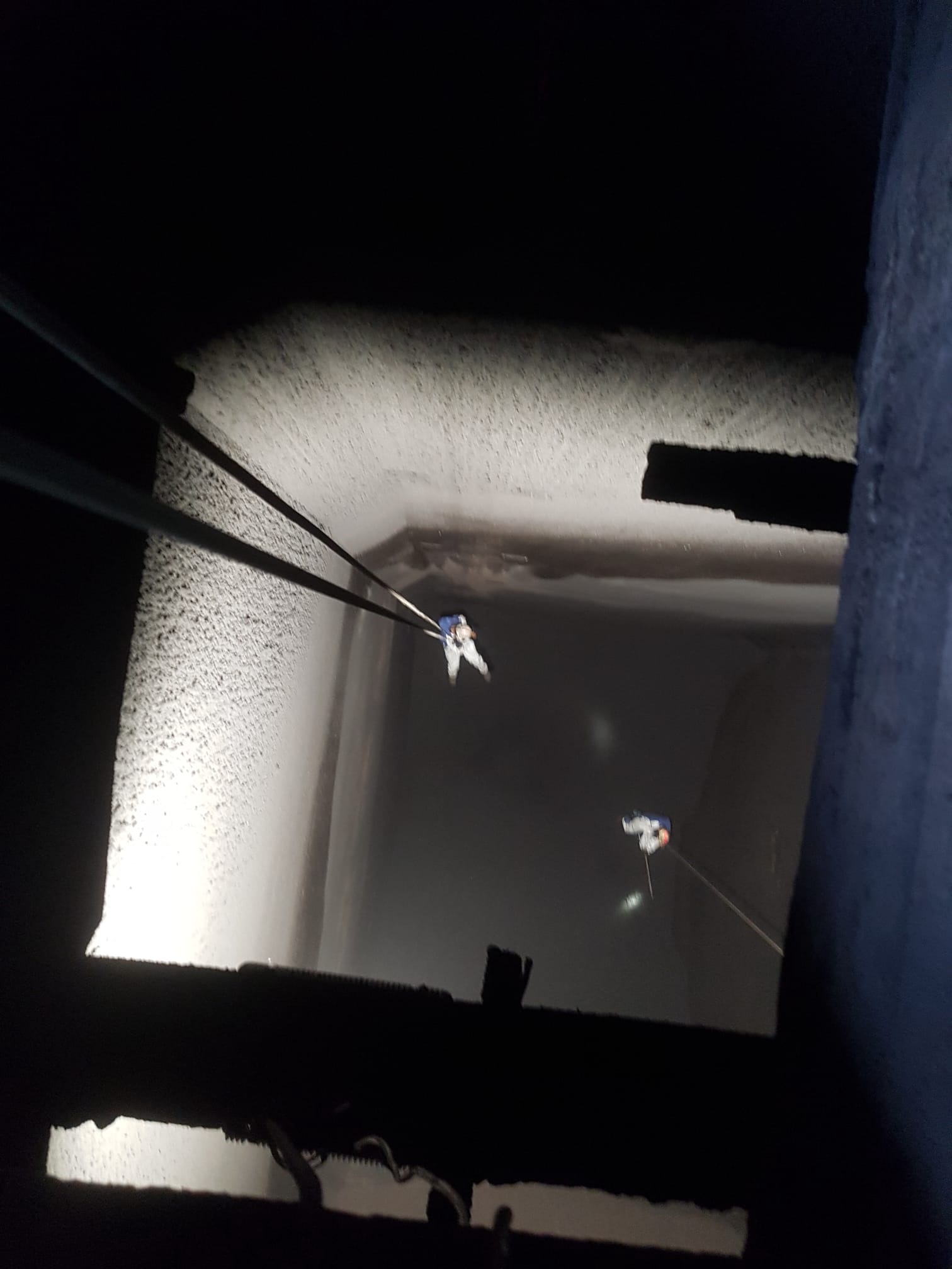 Dwóch alpinistów na linach wewnątrz ciemnego szybu, oświetlonych punktowo, widok z góry na dół, z elementami konstrukcji w kadrze.