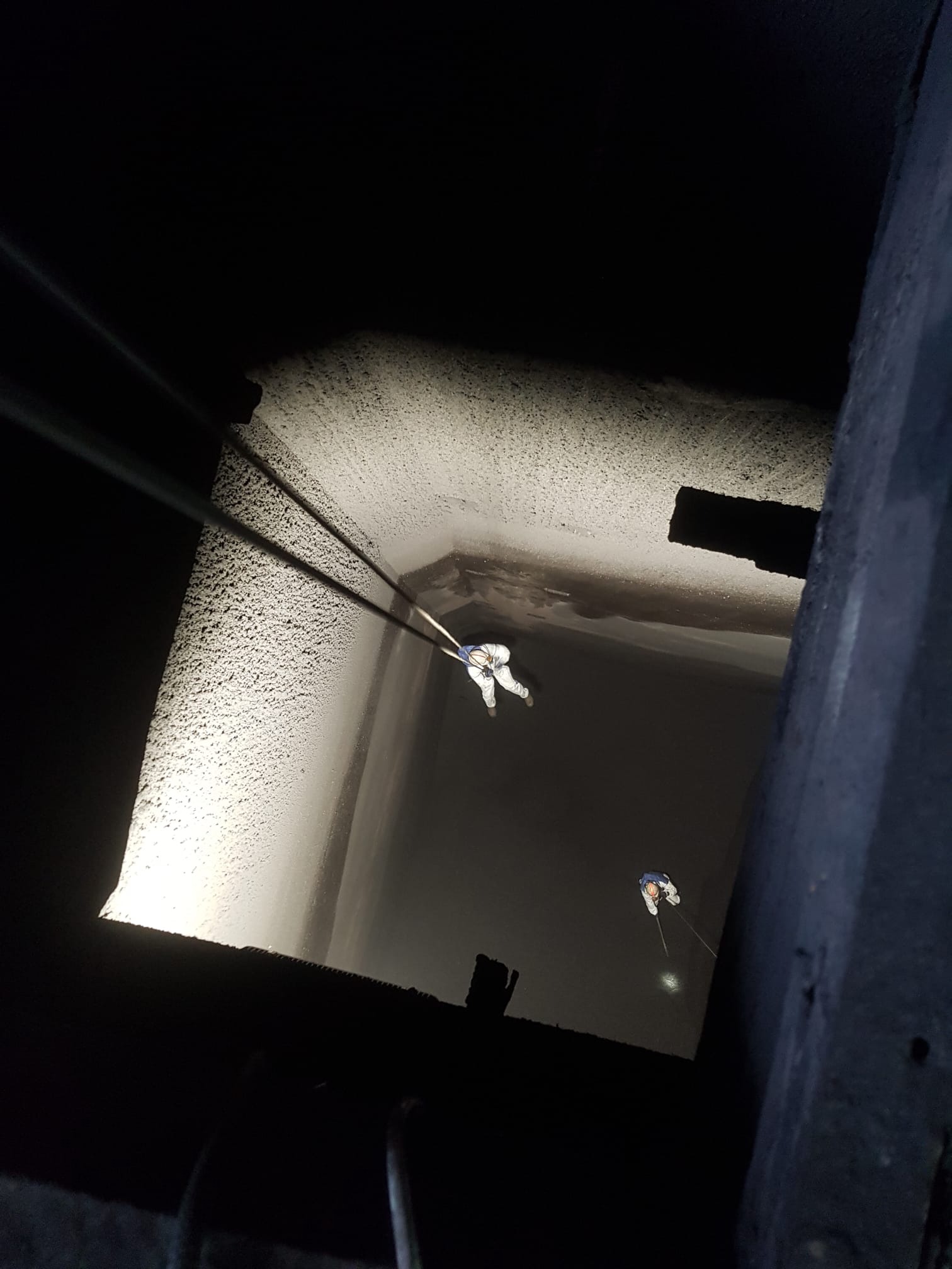 Dwóch pracowników na linach zabezpieczających, w kombinezonach, wykonuje prace na wysokości na ciemnej elewacji budynku. Ujęcie z dołu, z widocznymi linami.