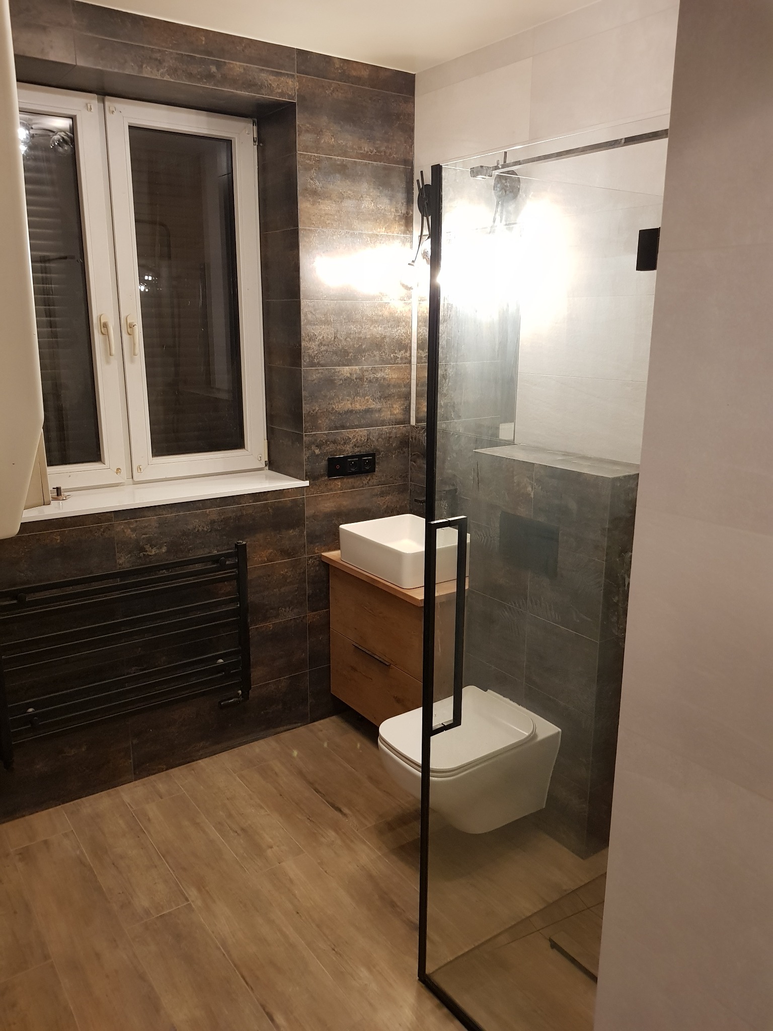 Nowoczesna łazienka z brązowymi płytkami imitującymi rdzę, kabiną prysznicową ze szkła hartowanego z czarnymi profilami, wiszącą toaletą i umywalką nablatową na drewnianej szafce.