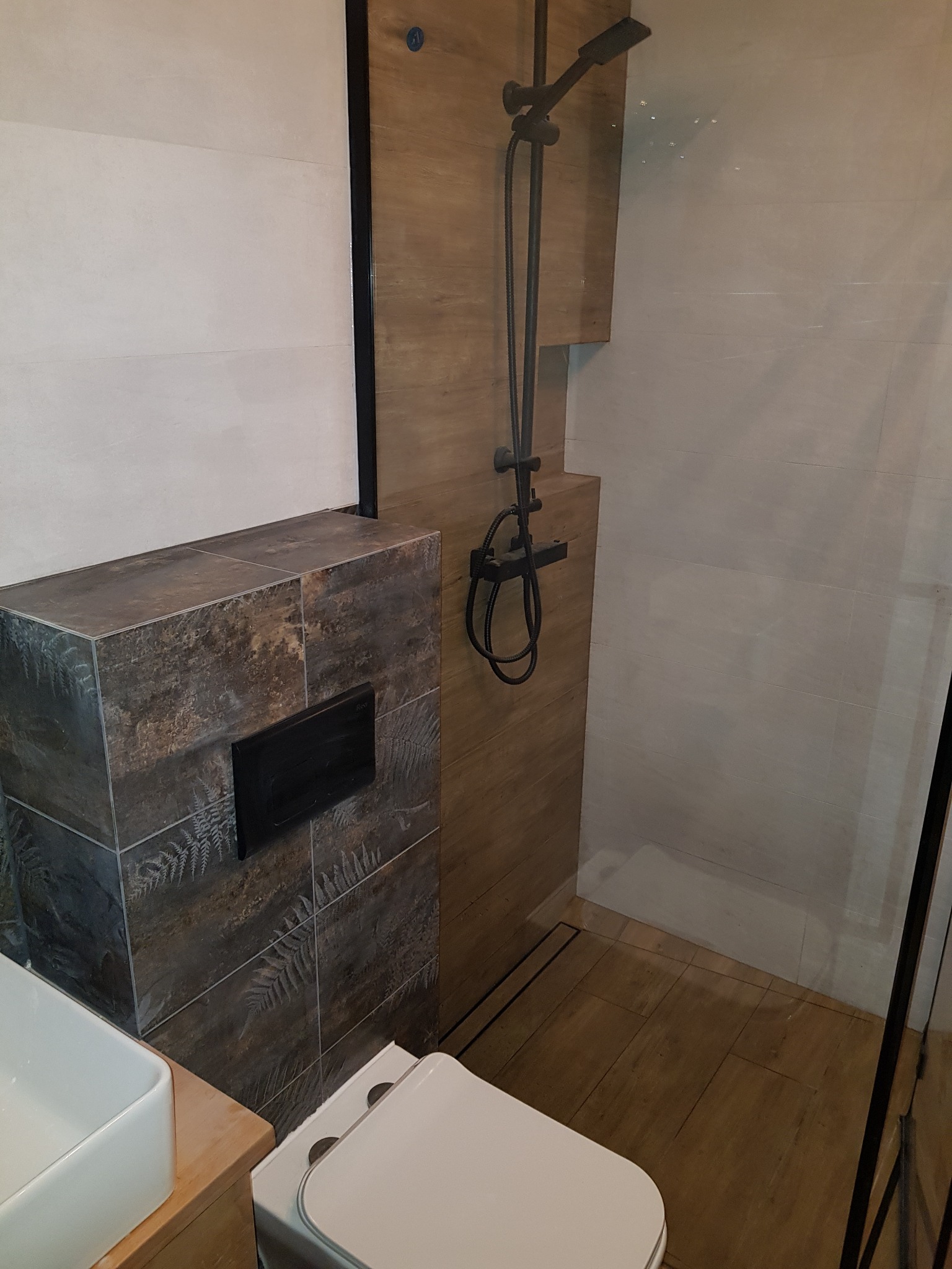 Nowoczesna łazienka z kabiną prysznicową typu walk-in, deszczownicą w kolorze czarnym, toaletą i umywalką, wykończona płytkami imitującymi drewno i kamień z motywem paproci.