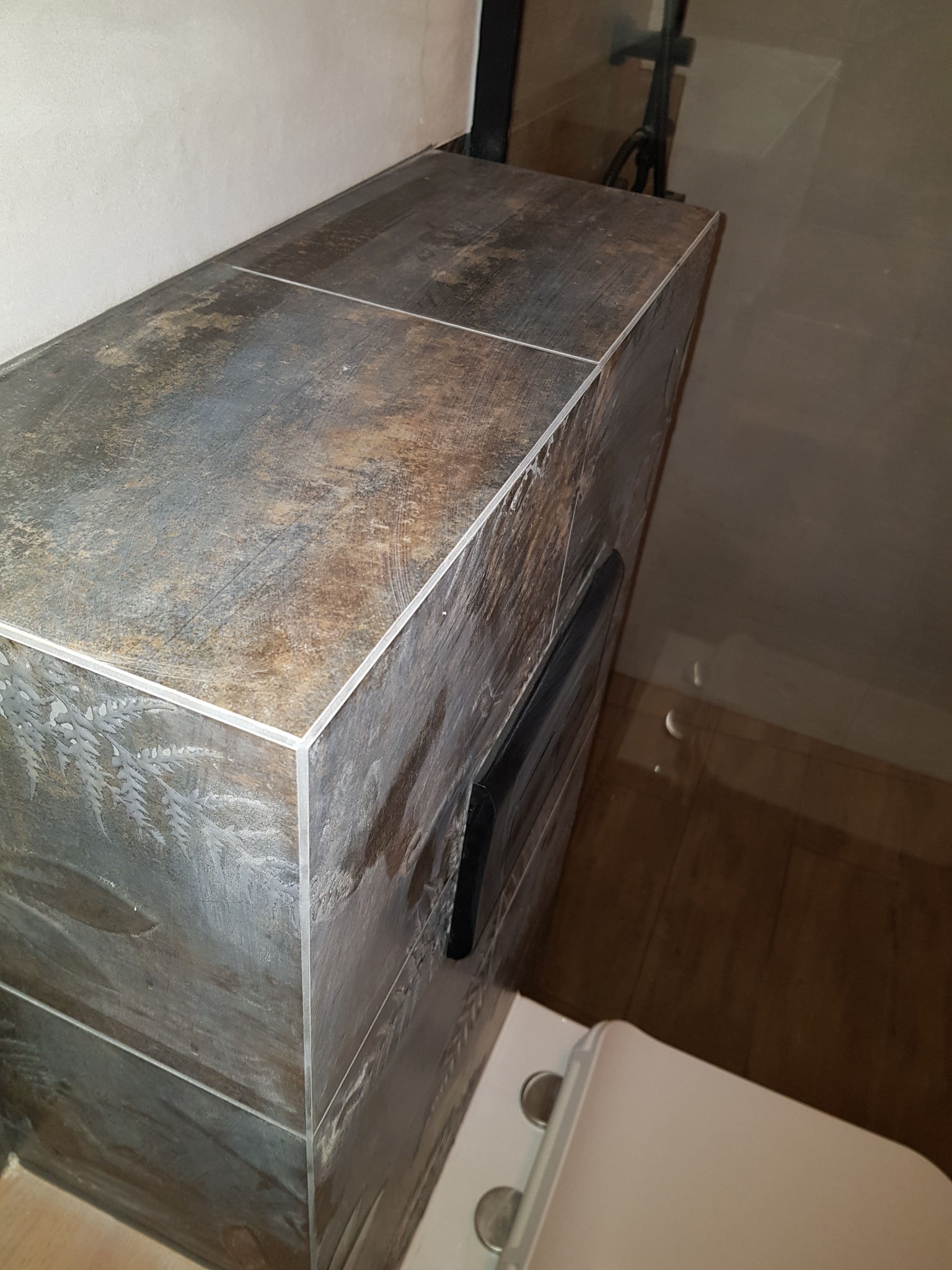 Obudowa toalety z płytek imitujących drewno z delikatnym motywem roślinnym, widoczny fragment toalety i bateria prysznicowa w tle.