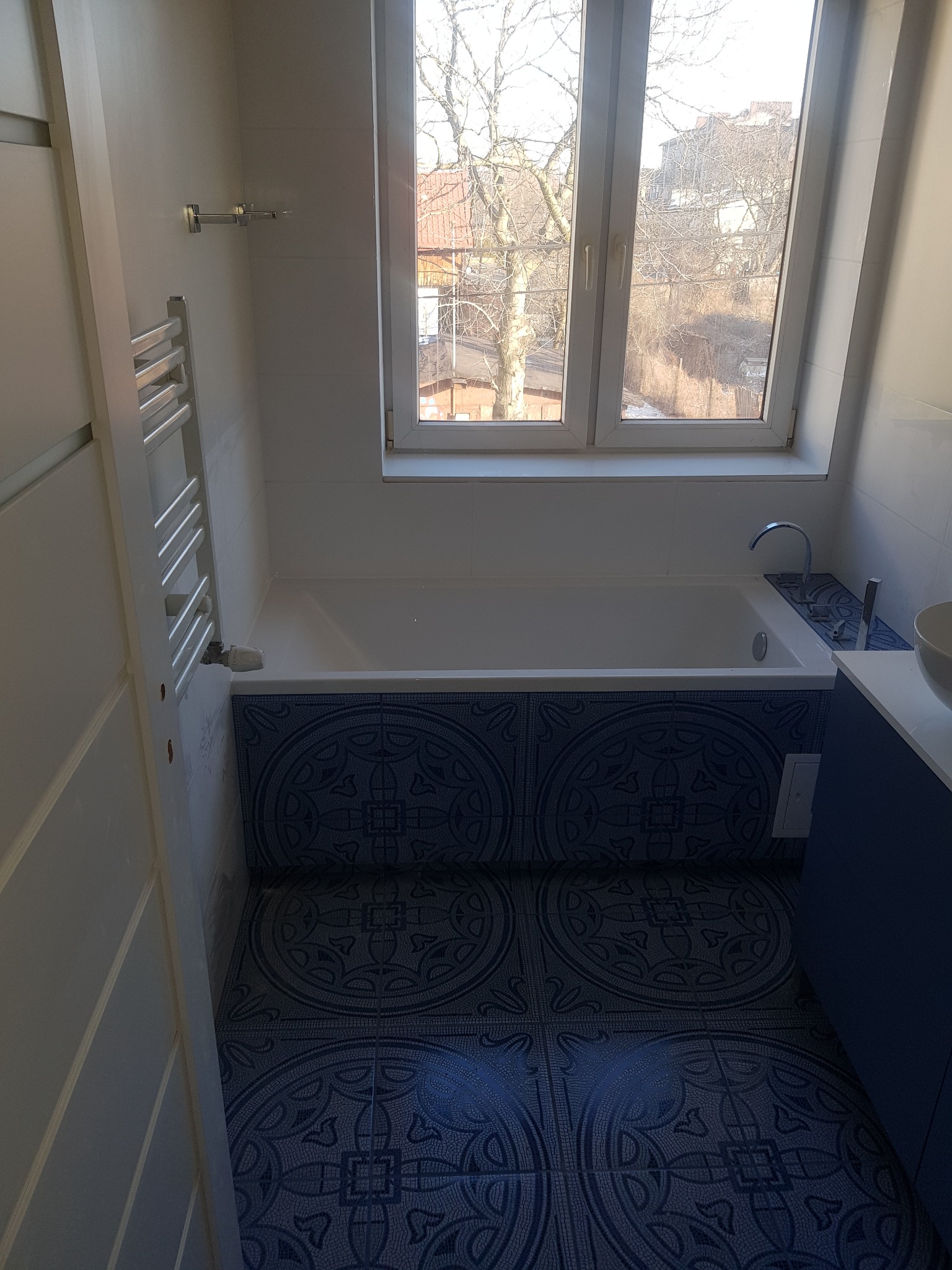 Wykończona łazienka z białą wanną, niebieskimi płytkami z motywem rozet na obudowie wanny i podłodze, grzejnikiem drabinkowym oraz oknem z widokiem na drzewa i budynki.