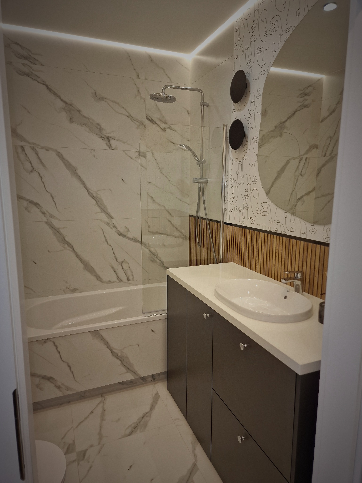 Nowoczesna łazienka z wanną obudowaną marmurowymi płytkami, chromowaną armaturą prysznicową, umywalką nablatową na szafce w kolorze ciemnego brązu oraz okrągłym lustrem z podświetleniem LED...