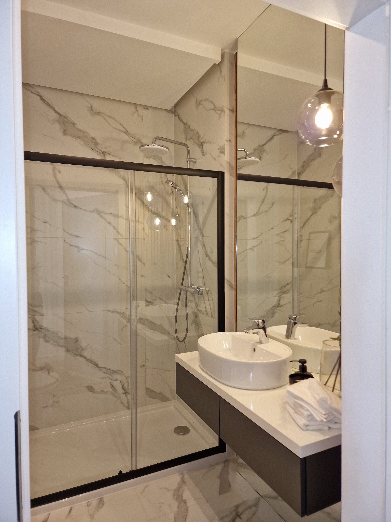 Nowoczesna łazienka z kabiną prysznicową typu walk-in z czarną ramą, umywalką nablatową na szafce w kolorze grafitowym i lustrem z odbiciem lampy wiszącej.