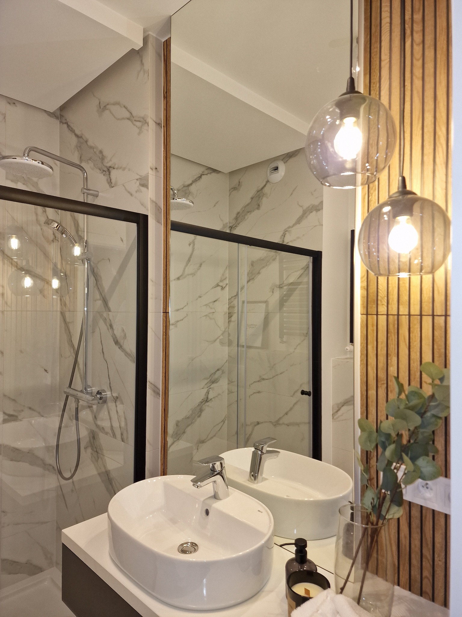 Nowoczesna łazienka z kabiną prysznicową typu walk-in, umywalką nablatową, lustrem i dekoracyjną ścianą z drewnianych paneli oraz wiszącymi szklanymi lampami.