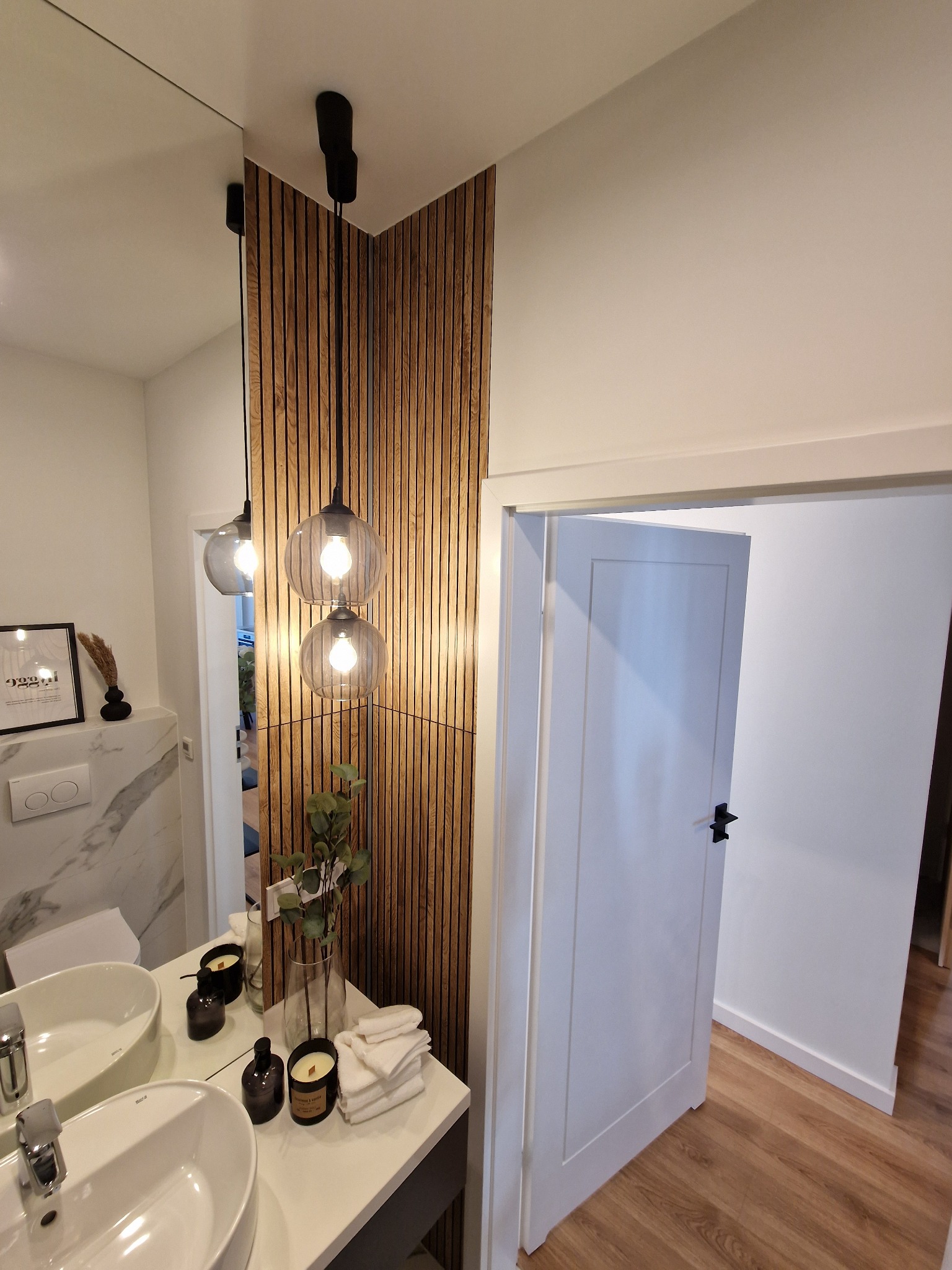 Nowoczesna łazienka we Wrocławiu z podwójną umywalką, drewnianymi panelami ściennymi i designerskimi lampami wiszącymi z żarówkami Edison, widok na otwarte drzwi.