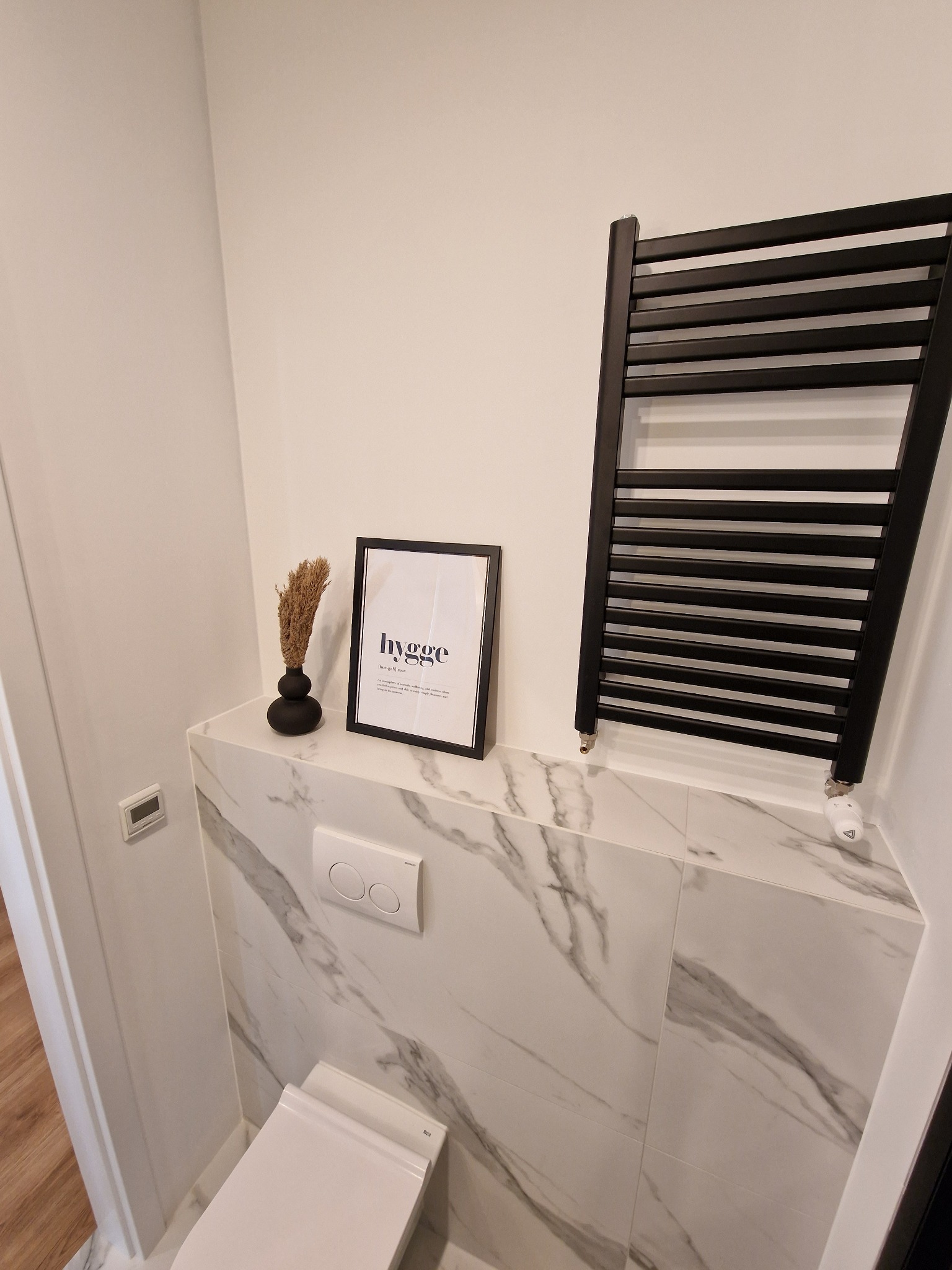 Nowoczesna toaleta z marmurowymi płytkami, czarnym grzejnikiem drabinkowym, dekoracją z napisem 'hygge' i czarnym wazonem z suszem.