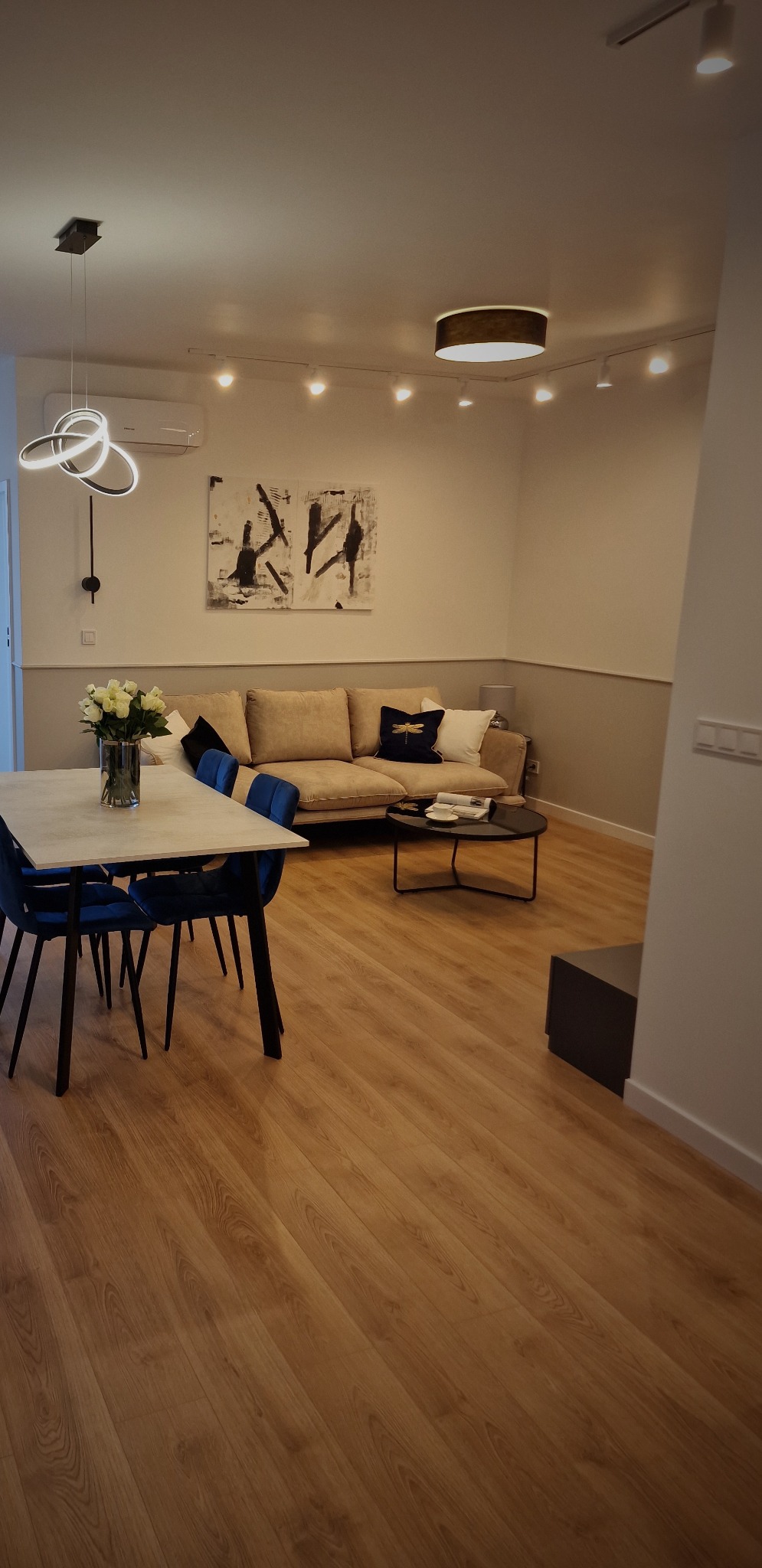 Jasne wnętrze salonu z drewnianą podłogą, beżową sofą z poduszkami, okrągłym stolikiem kawowym i stołem z niebieskimi krzesłami, oświetlone nowoczesnymi lampami sufitowymi.