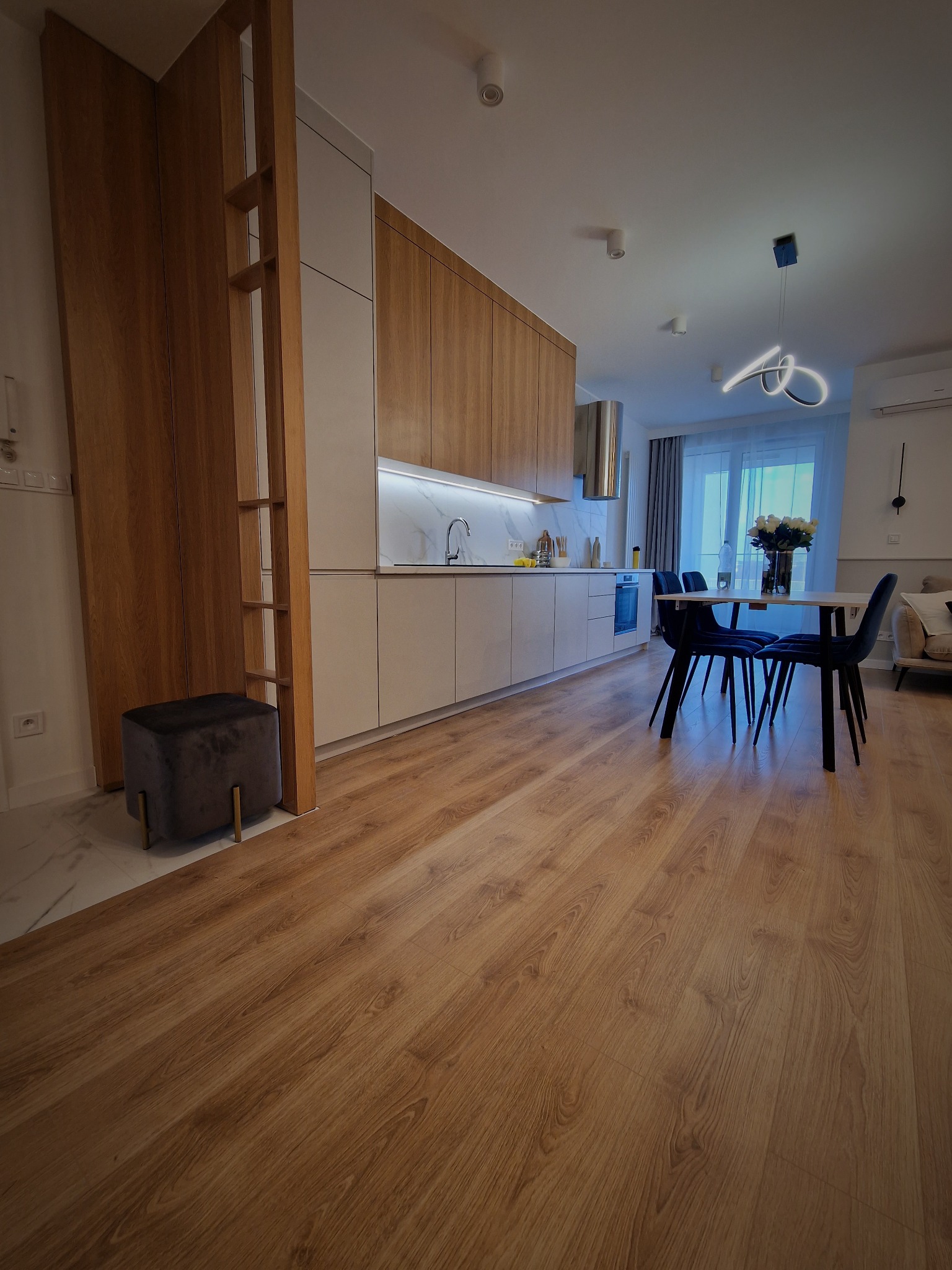 Nowoczesna kuchnia z drewnianymi frontami szafek, białym blatem i marmurowym backsplash, widok na stół z krzesłami i drewnianą podłogę.