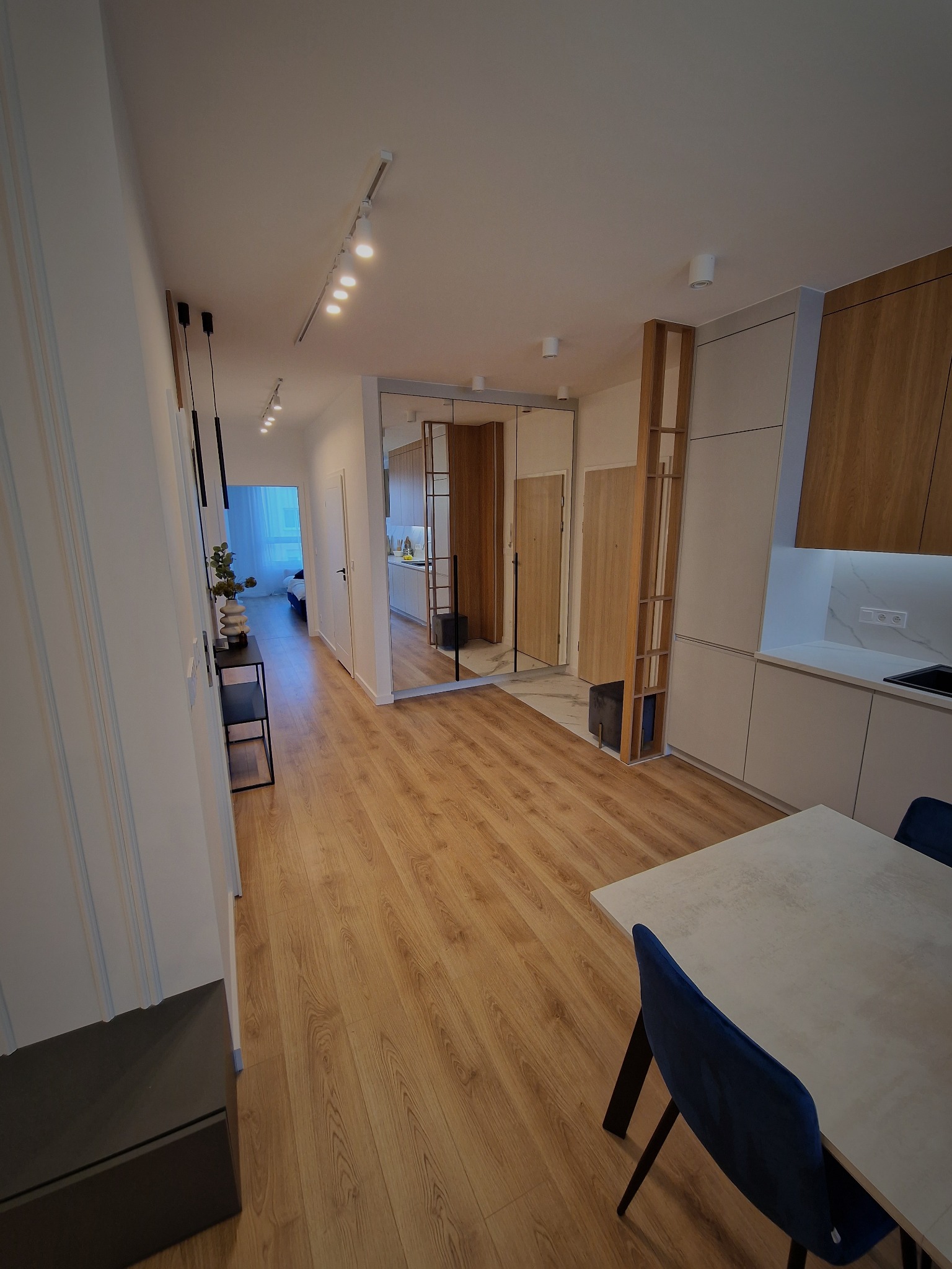 Wnętrze mieszkania po remoncie: widok na korytarz z elementami dekoracyjnymi, lustro powiększające przestrzeń, parkiet na podłodze i fragment kuchni z nowoczesnymi szafkami.