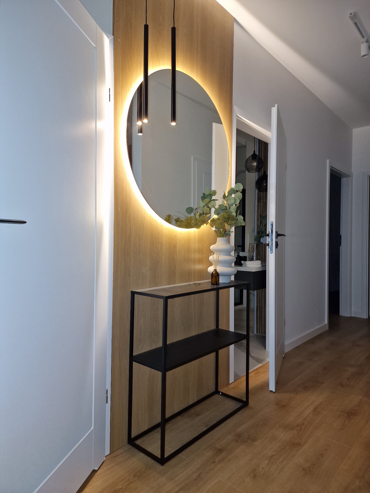 Nowoczesny przedpokój z okrągłym lustrem z podświetleniem LED, drewnianą ścianką, czarną konsolą i białymi drzwiami.