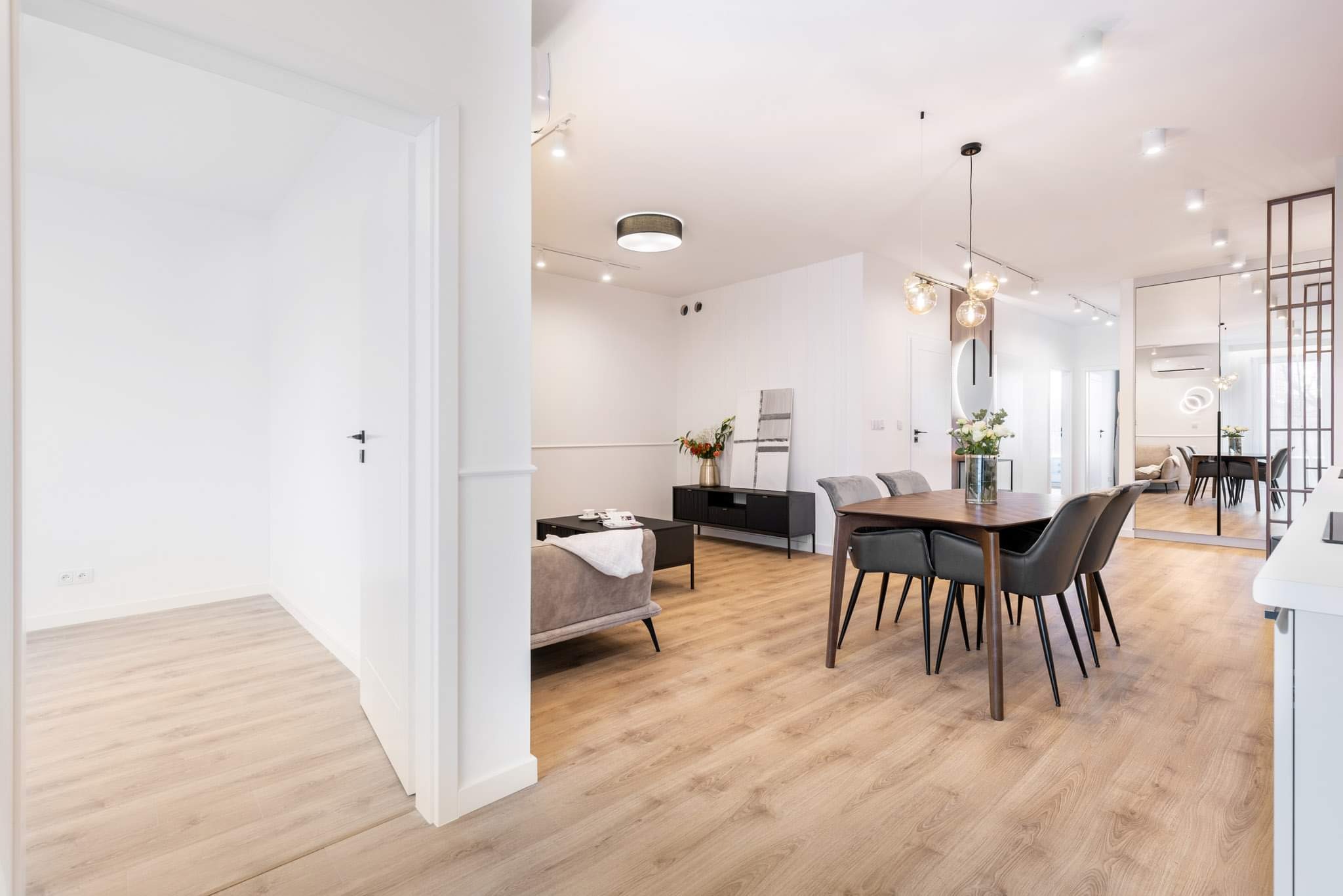 Jasne, przestronne wnętrze mieszkania z drewnianą podłogą, nowoczesnym oświetleniem i minimalistycznym umeblowaniem, widok z perspektywy wejścia do pokoju.