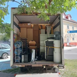 Wnętrze skrzyni ładunkowej wypełnione kartonami, krzesłami, komputerami i dystrybutorem wody, widoczne z rampy załadunkowej, z budynkiem Poczty Polskiej w tle.