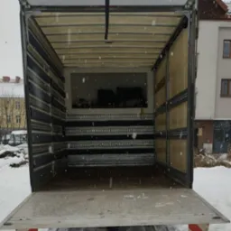 Pusta przestrzeń ładunkowa otwartego samochodu dostawczego widziana z tyłu, z widocznymi szynami mocującymi na ścianach i podłodze, na zewnątrz śnieg i budynek w tle.