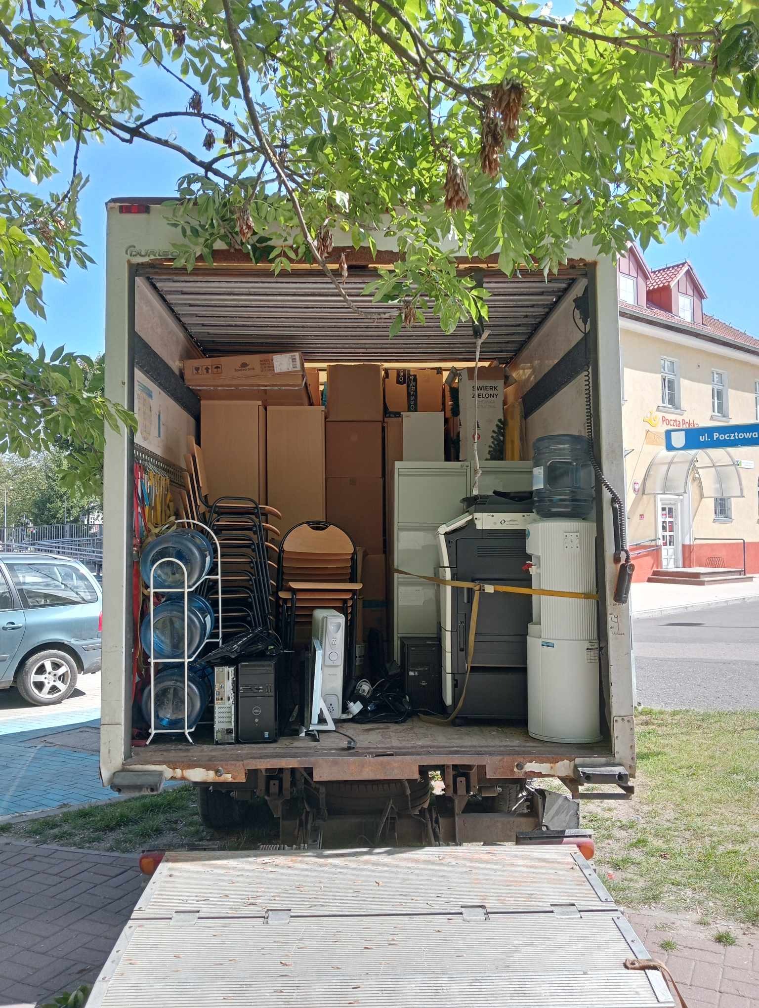 Wnętrze skrzyni ładunkowej wypełnione kartonami, krzesłami, komputerami i dystrybutorem wody, widoczne z rampy załadunkowej, z budynkiem Poczty Polskiej w tle.