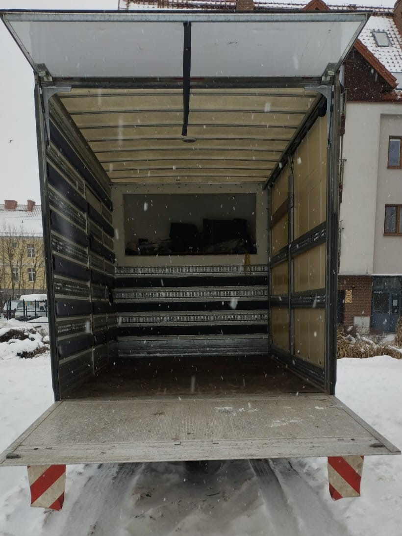 Pusta przestrzeń ładunkowa otwartego samochodu dostawczego widziana z tyłu, z widocznymi szynami mocującymi na ścianach i podłodze, na zewnątrz śnieg i budynek w tle.