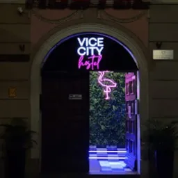 Wejście do hostelu Vice City z neonowym napisem i motywem flaminga, roślinna ściana w tle, podświetlane schody z szachownicą.
