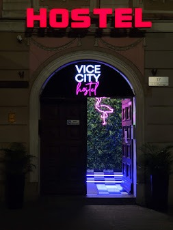 Wejście do hostelu Vice City z neonowym napisem i motywem flaminga, roślinna ściana w tle, podświetlane schody z szachownicą.