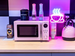 Różowa mikrofalówka na blacie kuchennym, obok czajnik w tym samym kolorze, neonowy napis z filiżanką kawy, puszka z napisem 'Herbalia' i słoik z kawą Gold.