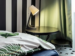 Nowoczesna lampka nocna z abażurem na okrągłym stoliku z metalu, obok łóżka z pościelą w liście, na tle ściany w czarno-białe pasy i zielonej zasłony.