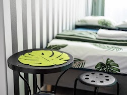 Pokój hotelowy z motywem roślinnym: stolik z zieloną podkładką w kształcie liścia monstery na tle łóżka z pościelą w liście, czarno-biała tapeta w pasy.