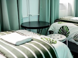 Pokój z dwoma łóżkami pojedynczymi, pościel w zielone paski i motywy liści, mały okrągły stolik nocny między łóżkami, turkusowe zasłony w tle.