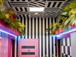 Korytarz w hotelu SPA z geometrycznym wzorem w czarno-białe pasy na ścianach i suficie, oświetlony neonowym różowym światłem, z roślinami tropikalnymi w donicach po bokach i różowymi drzwiami...