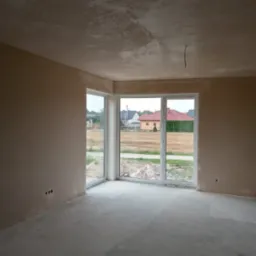 Surowe wnętrze po tynkowaniu maszynowym, widoczne niedoskonałości na suficie, okna PCV z widokiem na domy jednorodzinne, wystające przewody elektryczne.