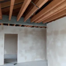 Wnętrze w trakcie prac wykończeniowych: surowy strop drewniany, ściany po wstępnym wyrównaniu, widoczna szara podłoga i otwór drzwiowy z leżącą miotłą.