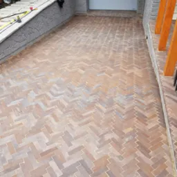 Ułożona kostka brukowa w jodełkę na podjeździe, widoczny fragment garażu i schod&oacute;w wejściowych, jasne spoiny między kostkami.