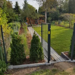 Widok na ogr&oacute;d z otwartą metalową furtką, świeżo posadzona trawa, ścieżka z jasnego żwiru, drewniana altana w tle.