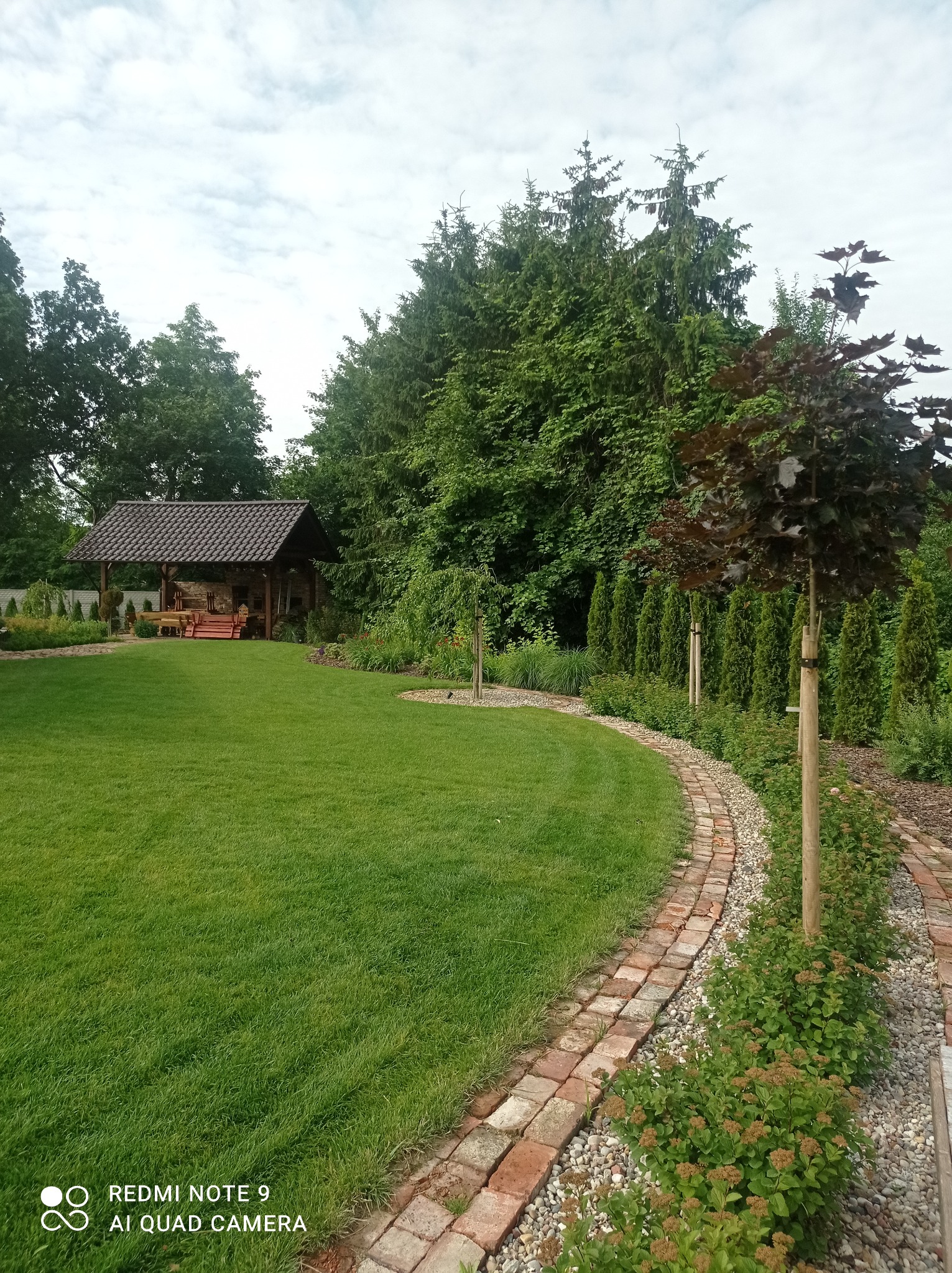 Zadbany ogród z równo przystrzyżonym trawnikiem, brukowaną ścieżką z czerwonej kostki, altaną w tle i drzewami, w tym jeden ozdobny klon o purpurowych liściach.