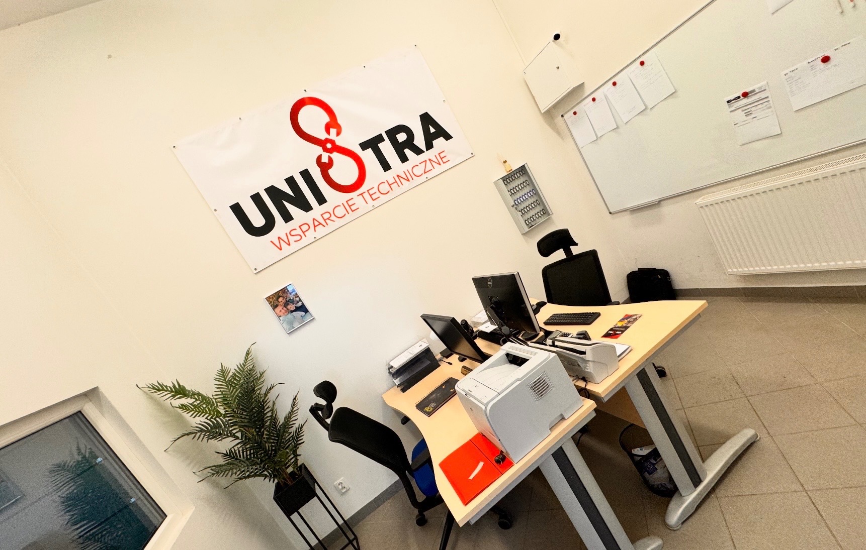 Biuro firmy wsparcia technicznego z logo UNIOTRA na ścianie. Widoczny biurko z komputerem, drukarką i rośliną doniczkową. Tablica suchościeralna w tle.