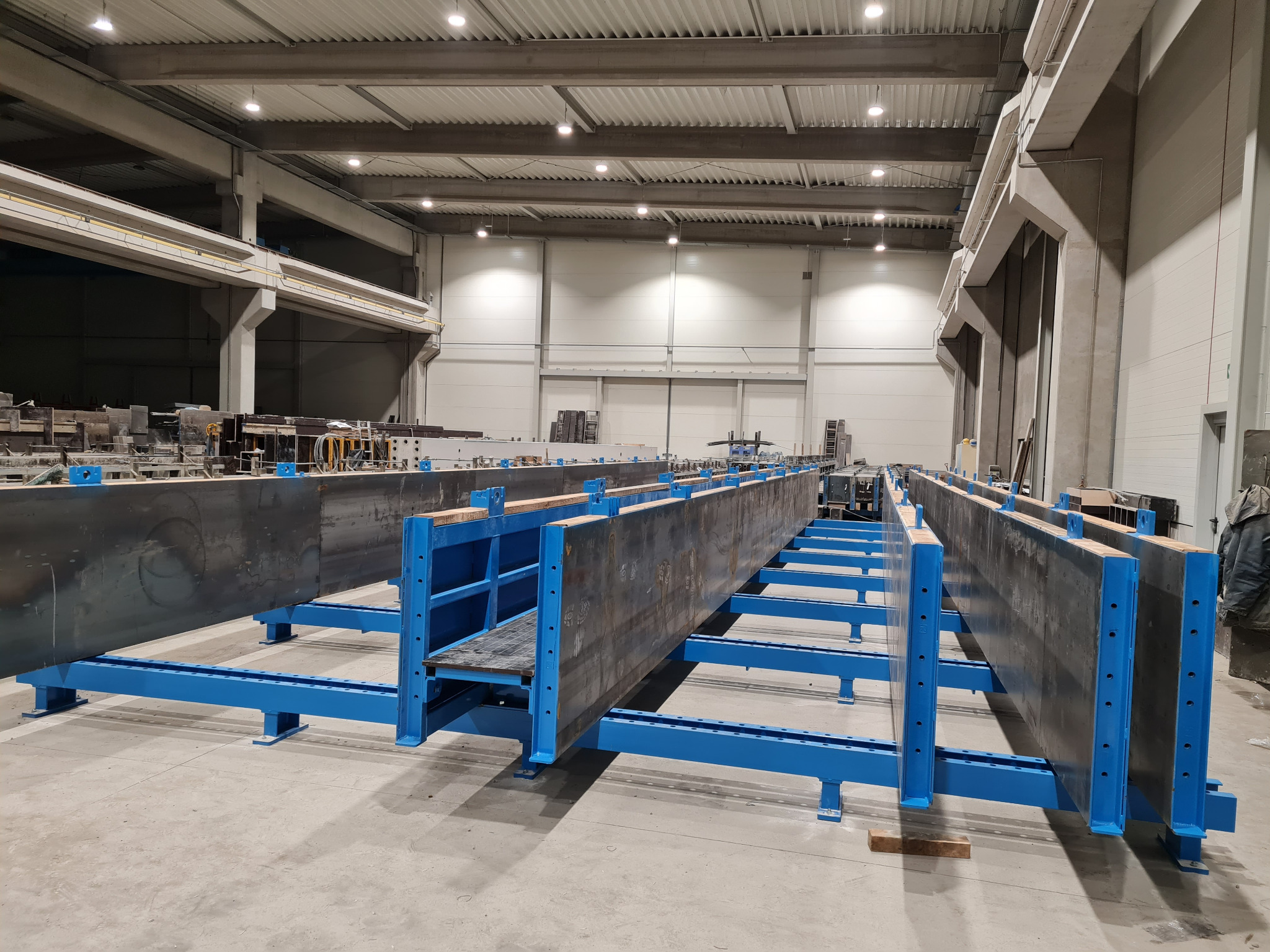 Wnętrze hali produkcyjnej z widocznymi stalowymi formami do odlewów betonowych, ustawionymi na niebieskich, metalowych konstrukcjach wsporczych.