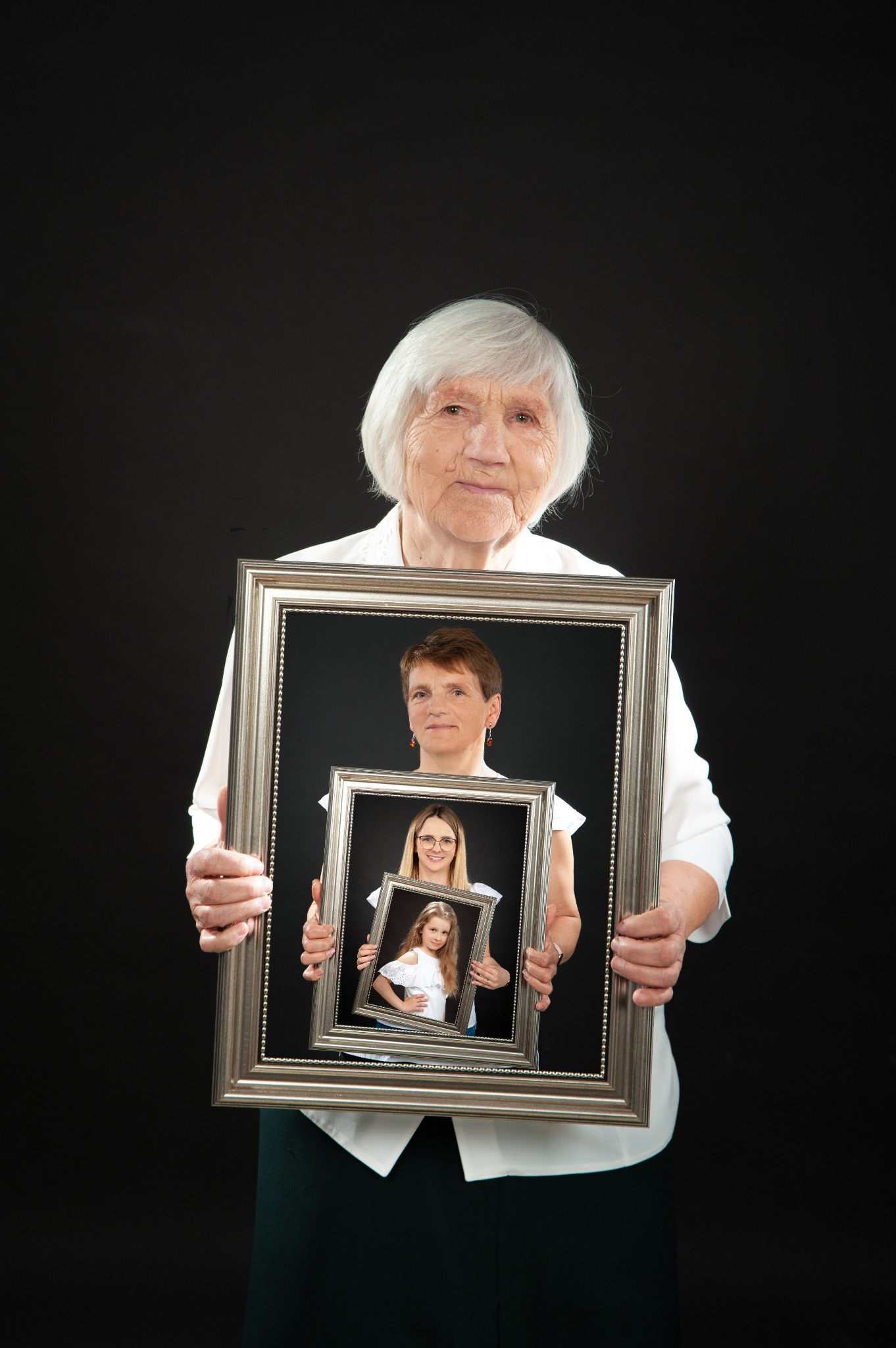 Starsza kobieta trzyma w rękach ramę ze zdjęciem córki, która trzyma ramę ze zdjęciem wnuczki, na czarnym tle.