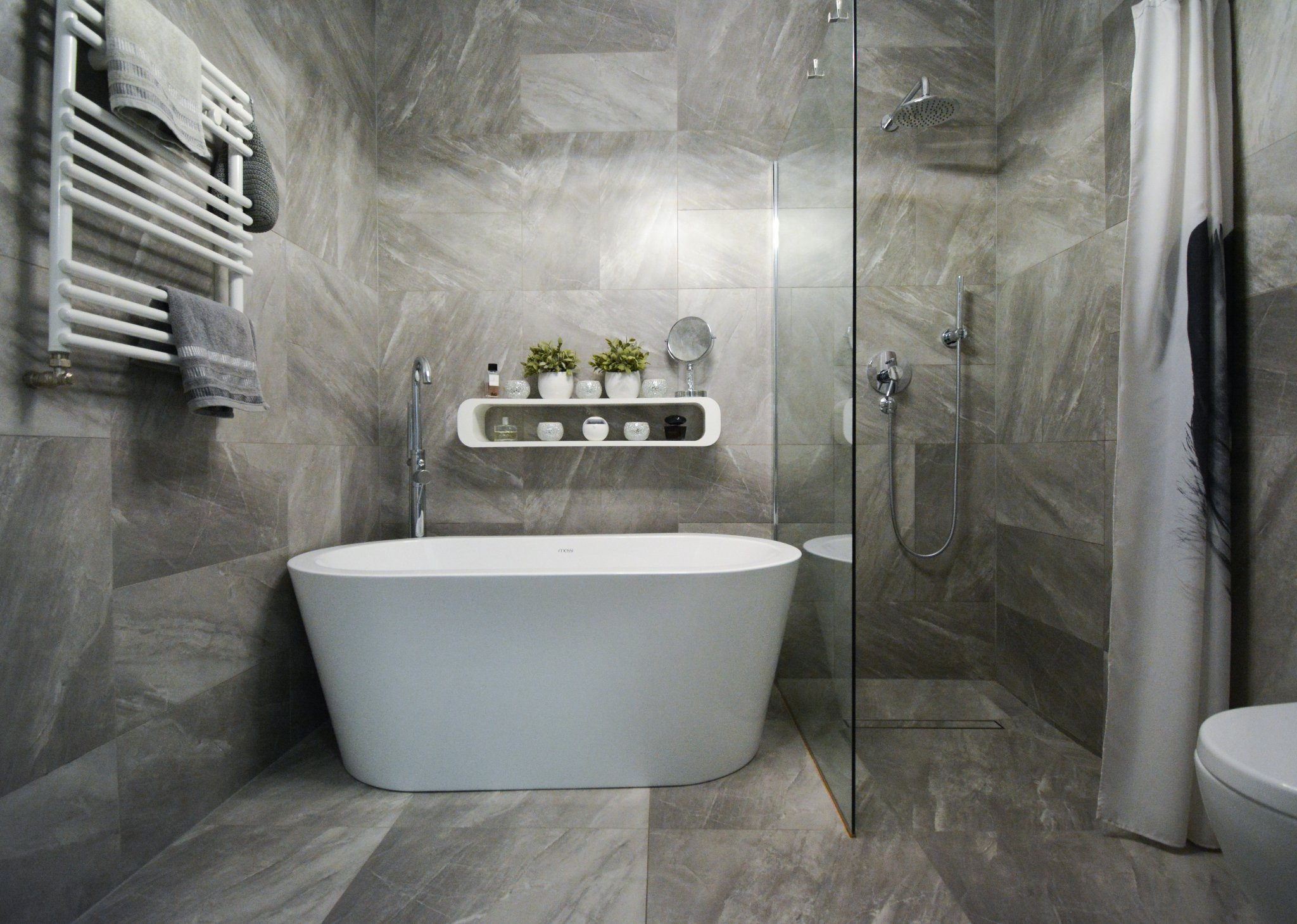 Nowoczesna łazienka z wolnostojącą białą wanną, szarymi płytkami imitującymi marmur, szklaną kabiną prysznicową i grzejnikiem drabinkowym z ręcznikami.