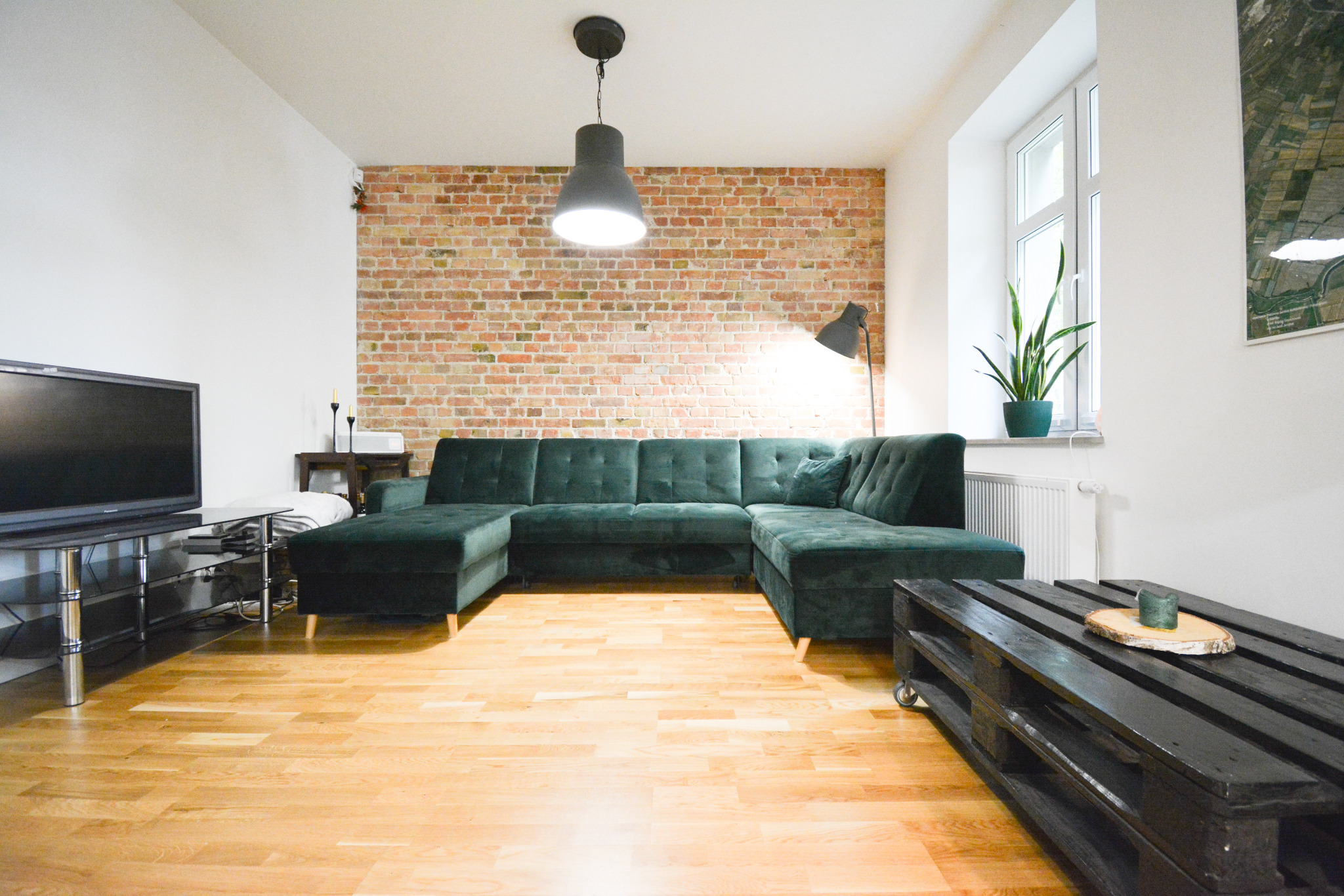 Salon w Gdańsku z ceglaną ścianą, ciemnozielonym narożnikiem, drewnianą podłogą i lampą wiszącą w stylu industrialnym.