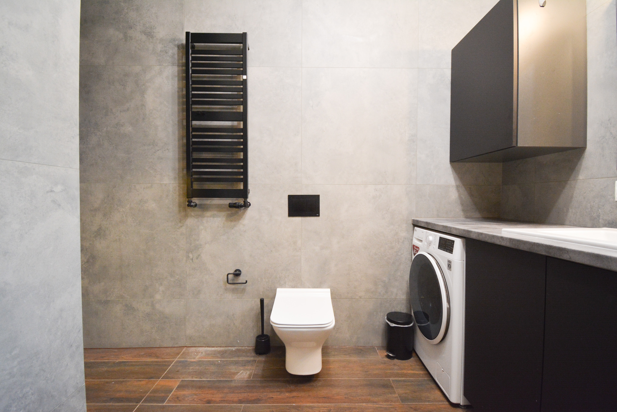 Nowoczesna łazienka z brązową drewnopodobną podłogą, szarą płytką na ścianach, wiszącą toaletą, czarnym grzejnikiem drabinkowym i pralką LG obok umywalki w szafce.