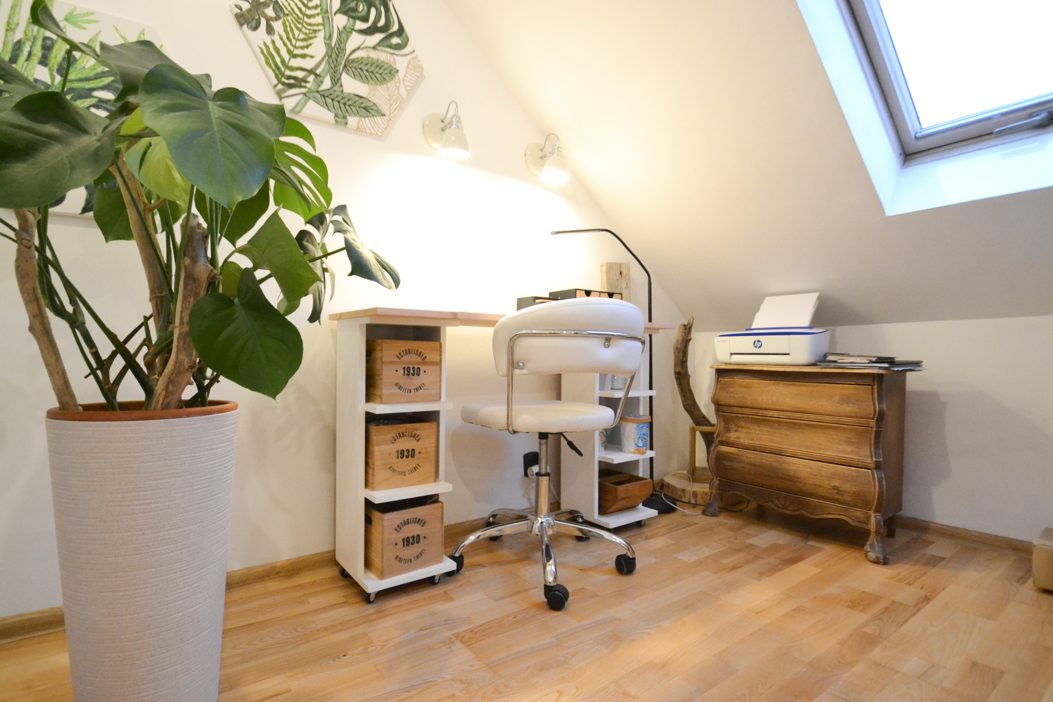 Jasne, domowe biuro na poddaszu z drewnianą podłogą, biurkiem z drewnianymi skrzynkami, białym krzesłem obrotowym, komodą z drukarką i dużą rośliną doniczkową.