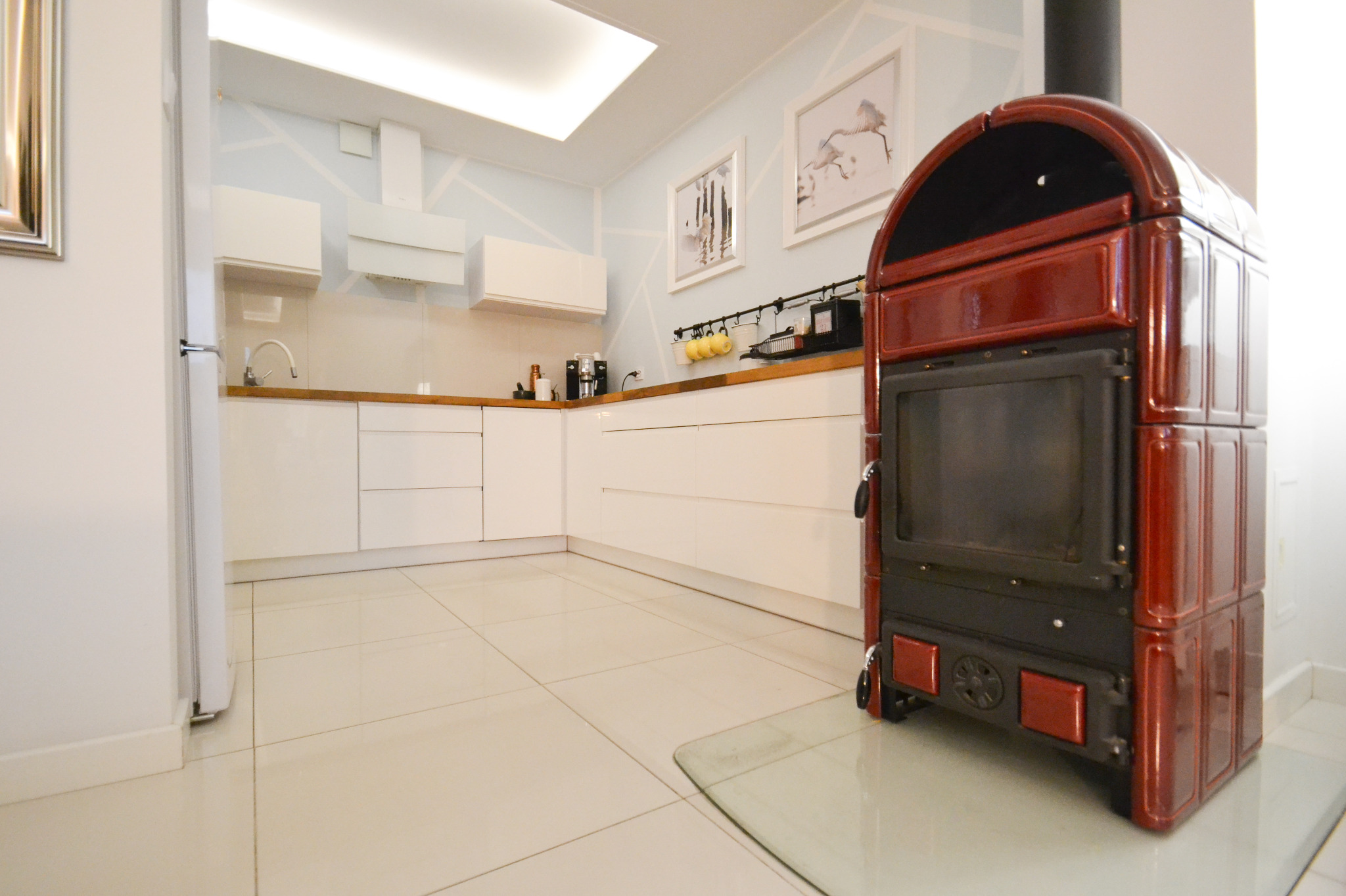 Nowoczesna biała kuchnia z drewnianym blatem i czerwonym piecem kaflowym na przeszklonej podstawie, widok z niskiej perspektywy.