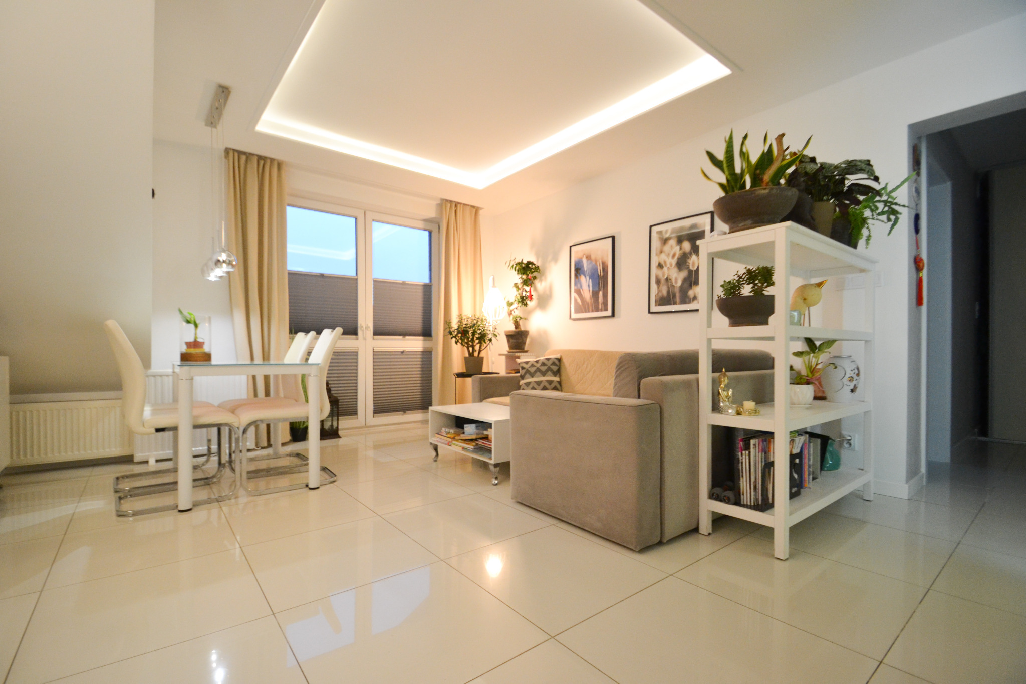 Jasny salon z beżową sofą, białym regałem z roślinami i dekoracjami, oraz stołem z krzesłami przy oknie z roletami.