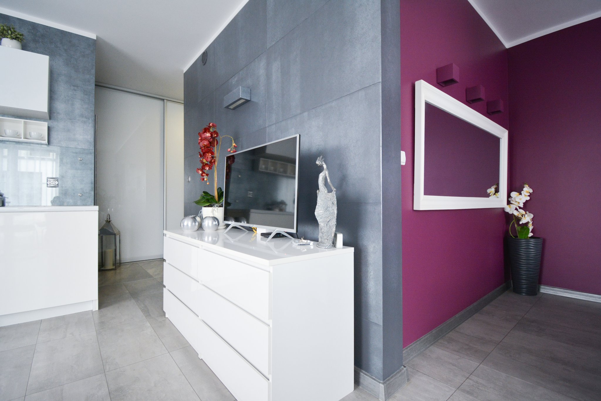 Nowoczesne wnętrze z szarą ścianą z płytek, białymi meblami, telewizorem, dekoracyjną figurką, fioletową ścianą z lustrem w białej ramie i kwiatami w wazonie.
