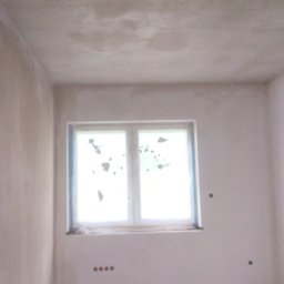 LISBUD TOMASZ LISTWAN - Surowe, jasne wnętrze pokoju po wstępnych pracach wykończeniowych, z oknem zabezpieczonym folią i ścianami pokrytymi szarym tynkiem, widoczne gniazdka elektryczne i nierówności przy podłodze.