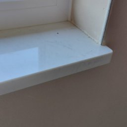 LISBUD TOMASZ LISTWAN - Biały parapet z PCV zamontowany w oknie, ściana pomalowana na beżowo, widoczne ślady po uszczelnianiu silikonem.