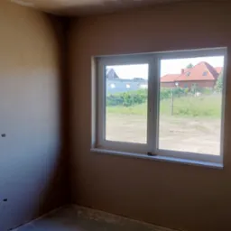 Pokój w stanie surowym z oknem, ściany pokryte świeżym tynkiem, widoczne puszki elektryczne bez osprzętu, za oknem widok na budowę i dom z czerwonym dachem.