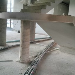 LisBud - Surowe betonowe schody w trakcie budowy, wsparte na okrągłych kolumnach, z widocznymi kablami i elementami konstrukcyjnymi na podłodze.