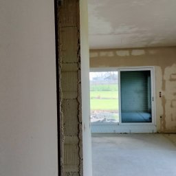 LisBud - Surowy stan ścian wewnątrz remontowanego pomieszczenia. Widoczne ślady po tynkowaniu i przygotowaniu do dalszych prac wykończeniowych, okno przesuwne na wprost.