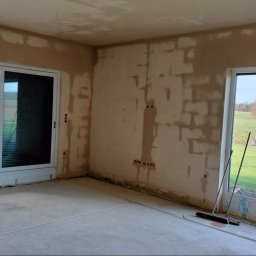 LISBUD TOMASZ LISTWAN - Wnętrze w trakcie tynkowania: ściany pokryte tynkiem, widoczne instalacje elektryczne, okna z widokiem na zewnątrz. Podłoga betonowa, przygotowana do dalszych prac wykończeniowych.