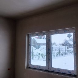 LISBUD TOMASZ LISTWAN - Pokój w trakcie prac wykończeniowych: świeżo położone gładzie na ścianach i suficie, białe okno z widokiem na zimowy krajobraz za oknem. Widoczne detale wykończenia.